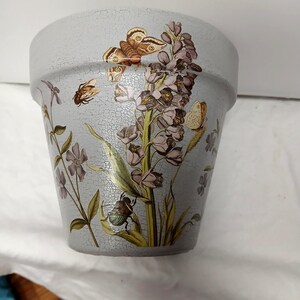 May include: A light gray flower pot with a crackle finish, decorated with a botanical illustration featuring a tall purple flower stalk, a brown butterfly, a small yellow butterfly, a flying insect, and a green beetle on the lower leaves.