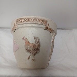 May include: Cream-coloured farmhouse-style ceramic pot with embossed details. Features a pig, a chicken, and the word "FARMHOUSE" across the top. The pot has a textured surface and is suitable for plants.