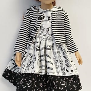 May include: A doll with auburn hair wearing a black and white striped cardigan over a dress featuring a musical staff and note print. The dress has a white bodice and a black skirt with white musical notes. It wears striped socks and white shoes.