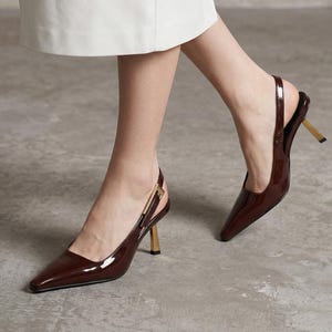 May include: Burgundy patent leather slingback heels with pointed toes and gold-coloured heels. The shoes have an ankle strap and a small gold buckle. The heels are approximately 7.6 cm high.