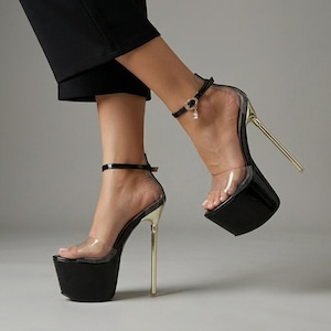 May include: Black platform high heels featuring a clear transparent strap across the toes, a thin ankle strap with a small dangling charm, and a metallic gold stiletto heel. The platform base is solid black, providing a stark contrast to the gold heel.