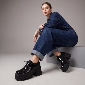 Pode incluir: Sapatos de plataforma em verniz preto com design de atacadores. Os sapatos t&ecirc;m sola grossa e de rasto e um salto alto e largo. O cal&ccedil;ado &eacute; usado com um macac&atilde;o de ganga.