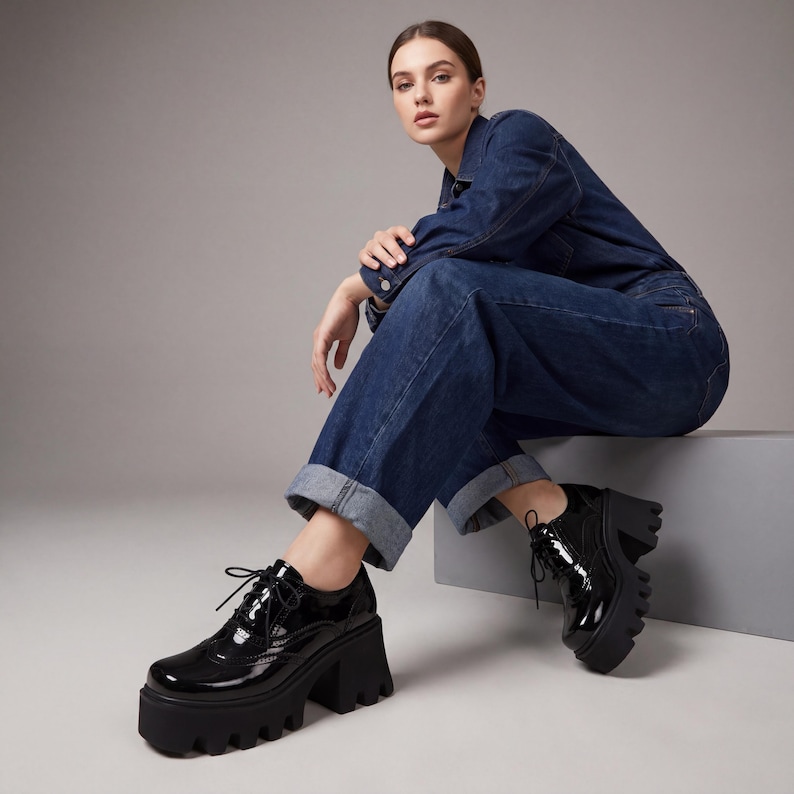 Pode incluir: Sapatos de plataforma em verniz preto com design de atacadores. Os sapatos t&ecirc;m sola grossa e de rasto e um salto alto e largo. O cal&ccedil;ado &eacute; usado com um macac&atilde;o de ganga.