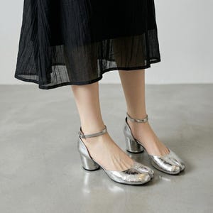 May include: Silver metallic heeled sandals with a Tabi toe design. The sandals have a chunky heel and an ankle strap with a buckle closure. The shoes are a shiny silver color.