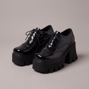 Pode incluir: Um par de sapatos oxford pretos com plataforma e acabamento brilhante. Os sapatos apresentam um design com atacadores, perfura&ccedil;&otilde;es decorativas e uma sola grossa e com rasto. Os saltos t&ecirc;m cerca de 10 cm de altura.