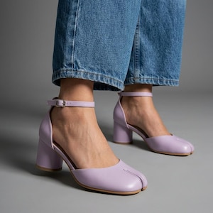 Könnte beinhalten: Lavendelfarbene Schuhe mit Blockabsatz und geteilter Zehenpartie, Knöchelriemen und Schnallenverschluss, kombiniert mit blauen Jeans. Die Schuhe zeichnen sich durch ein markantes Schlitzdesign an der Zehenkappe, einen stabilen Absatz und eine glatte Oberfläche aus.