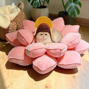 May include: A light-colored cat wearing a brown crocheted hat with a yellow semi-circular brim, resting in the center of a large, plush, pink flower-shaped cushion with multiple rounded petals arranged in a circular pattern.