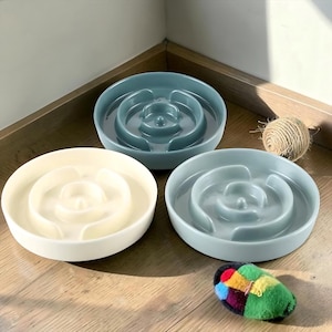 May include: Three round slow feeder bowls with internal raised ridges. One bowl is cream-colored, while the other two are a muted blue-gray. The bowls feature a smooth, glossy finish and a circular design intended to slow down eating speed.