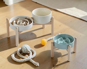 May include: A double elevated pet bowl set in white with a slow feeder insert and a separate single elevated pet bowl in light blue with a slow feeder insert, both featuring light wood legs, accompanied by a braided rope toy and a yellow ball.