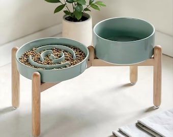 Pode incluir: Uma estação de alimentação dupla para animais de estimação com duas tigelas de cerâmica verde sálvia num suporte de madeira clara. Uma tigela contém ração seca e tem um design de labirinto em espiral, a outra é lisa e aberta.
