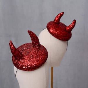 Puede incluir: Dos diademas de cuernos de diablo rojos con lentejuelas. Cada cuerno es curvo y está unido a una pequeña base redonda. Las diademas se exhiben en cabezas de maniquí blancas sobre un fondo gris.