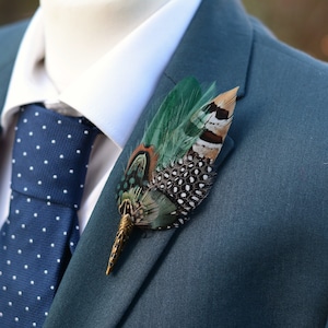 May include: A feather boutonniere with green, brown, and white feathers, attached to a teal suit jacket. The jacket is paired with a white dress shirt and a navy blue tie with white polka dots. The boutonniere has a gold-colored metal base.