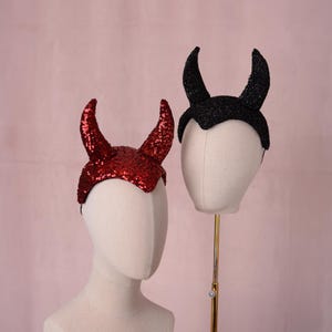 Può includere: Due cerchietti con corna da diavolo, uno ricoperto di paillettes rosse lucide e l'altro di glitter neri. Ogni cerchietto è esposto su una testa di manichino di colore neutro, mettendo in risalto il design a punta delle corna e la superficie.