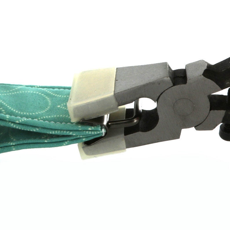 May include: Close-up of a black and gray tool with a white plastic jaw. The tool is holding a piece of teal fabric with a white pattern.