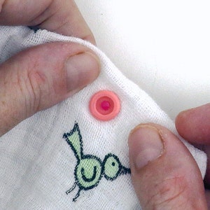 May include: A pink snap button with a white center is attached to a white fabric with a green bird design.