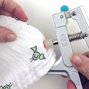 May include: A close-up of a silver snap press tool being used to attach a snap to a white fabric with a green bird design. The snap is pink and white.