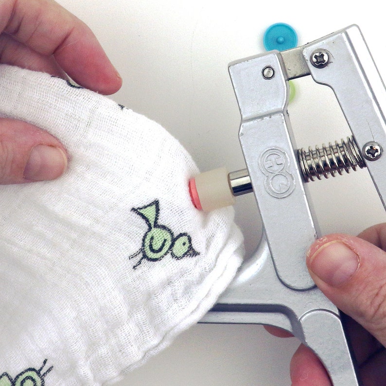 May include: A close-up of a silver snap press tool being used to attach a snap to a white fabric with a green bird design. The snap is pink and the tool has a spring mechanism.