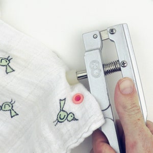 May include: A silver snap press tool being used to attach a pink snap to a white fabric with a green and black bird design.