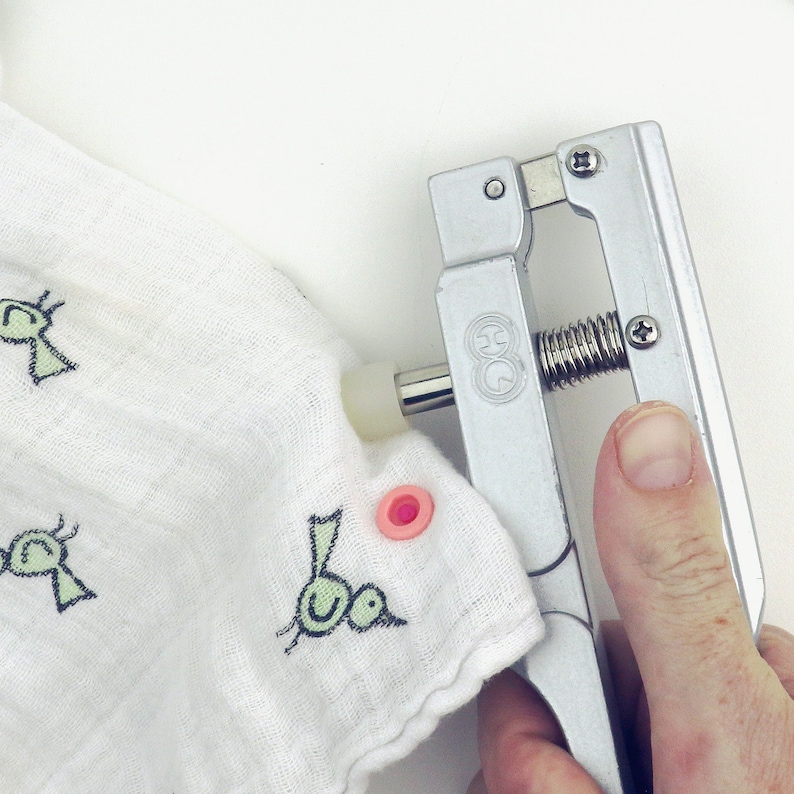 May include: A silver snap press tool being used to attach a pink snap to a white fabric with a green and black bird design.