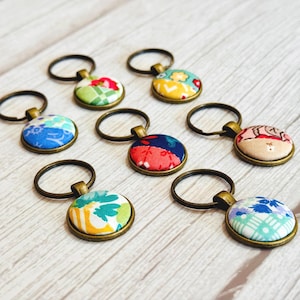 May include: A collection of seven circular keychains featuring fabric-covered buttons in various colorful patterns. Each button is set within a bronze-toned metal frame and attached to a matching bronze-toned metal split ring for keys.