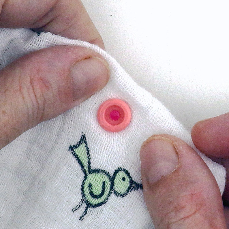 May include: A pink snap button with a white center is attached to a white fabric with a green bird design.