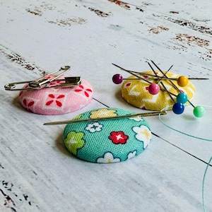 May include: Three colorful pin cushions with floral patterns. One pink cushion has red flower accents and a safety pin. A yellow cushion holds several pins with colorful heads. A teal cushion has white, red, and green floral accents.