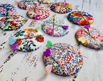 May include: A collection of round fabric pincushions featuring various floral patterns in vibrant colors. Each pincushion is pierced by two long sewing pins with heart-shaped heads in pastel shades of pink, blue, purple, yellow, and green.