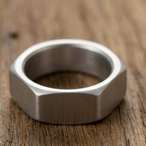 Puede incluir: Un anillo de metal plateado con forma de tuerca hexagonal. La banda presenta un acabado cepillado con lados planos y angulares que imitan la apariencia de los herrajes industriales. El interior del anillo es liso y de forma circular.
