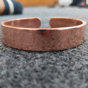 Pode incluir: Uma pulseira de punho ajustável na cor cobre. A pulseira tem um design plano e retangular com uma superfície texturizada. O design aberto permite um ajuste fácil. A pulseira tem uma cor castanho-avermelhada quente.
