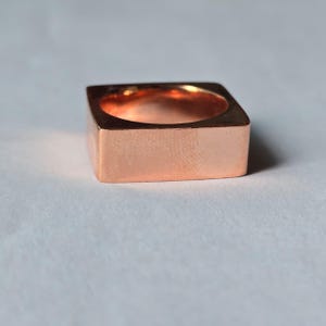 Op de afbeelding: Een vierkante koperen ring met een gepolijste afwerking. De ring heeft een holle binnenkant en een minimalistisch ontwerp. De afmetingen van de ring zijn ongeveer 1,9 cm breed en 1,9 cm hoog.
