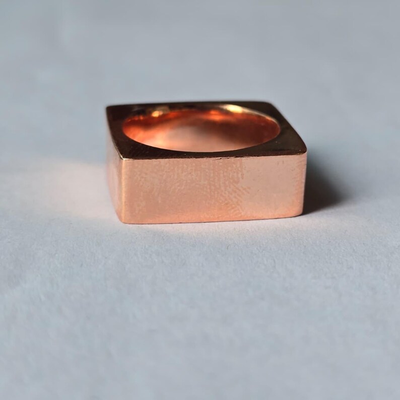 Op de afbeelding: Een vierkante koperen ring met een gepolijste afwerking. De ring heeft een holle binnenkant en een minimalistisch ontwerp. De afmetingen van de ring zijn ongeveer 1,9 cm breed en 1,9 cm hoog.
