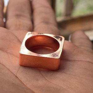 May include: A square copper ring with rounded inner edges. The ring has a smooth, polished finish and a hollow centre. The ring is a warm, reddish-brown colour and appears to be handmade. The ring is shown on a person's palm.