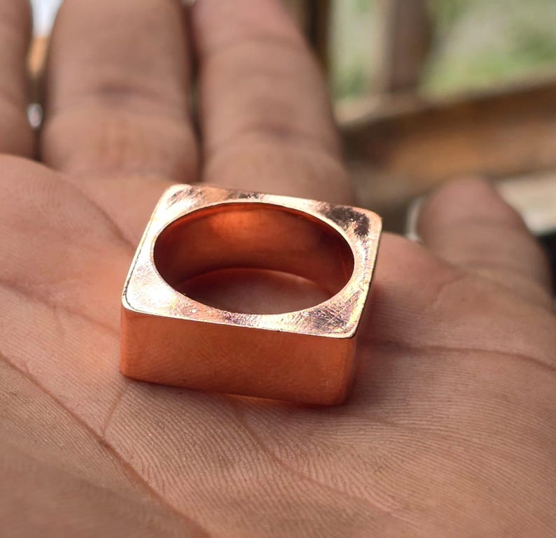 Op de afbeelding: Een vierkante koperen ring met afgeronde binnenranden. De ring heeft een gladde, gepolijste afwerking en een hol midden. De ring heeft een warme, roodbruine kleur en lijkt handgemaakt te zijn. De ring wordt op de handpalm van een persoon getoond.