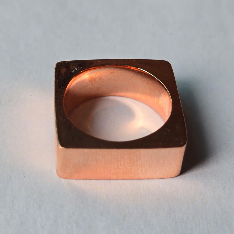 Op de afbeelding: Een vierkante koperen ring met een gepolijste afwerking. De ring heeft een holle binnenkant en een minimalistisch ontwerp. De koperkleur is warm en uitnodigend, waardoor het een veelzijdig accessoire is.