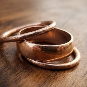 May include: A set of three copper-coloured rings stacked together. One ring features a wide, smooth, polished band, while the other two rings are thinner with a textured, hammered finish. The metal has a warm, metallic reddish-brown tone.