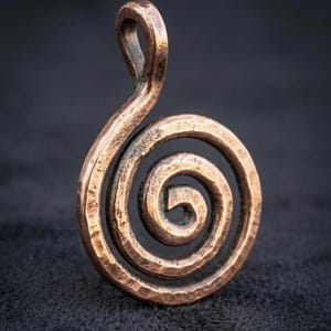 Może przedstawiać: Miedziany metalowy wisiorek z ręcznie kutym wzorem spirali. Wisiorek ma teksturowaną, rustykalną powierzchnię z wyraźną pętlą u góry do zawieszenia, prezentującą okrągły, zwinięty kształt z ciepłym, metalicznym wykończeniem.