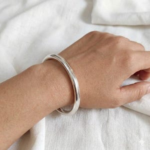 May include: A polished, silver bangle bracelet. The bracelet is a thick, rounded band. The jewellery is worn on a person's wrist. The background is a white, textured fabric.
