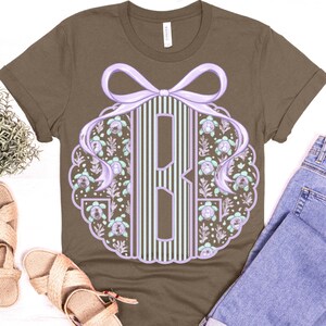 May include: Brown t-shirt with a monogram design in light purple, surrounded by a floral pattern and a bow. The shirt is paired with blue jeans and sandals. The design is centered on the shirt.