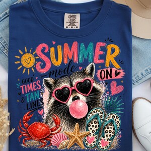 Könnte beinhalten: Marineblaues T-Shirt mit einem farbenfrohen Sommer-Design. Das Design zeigt einen Waschbären mit herzförmiger Sonnenbrille, eine Blase und Strandelemente. Der Text lautet "SUMMER mode ON".