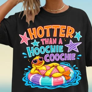 Peut inclure: T-shirt noir avec l'inscription "HOTTER THAN A HOOCHIE COOCHIE" en orange, bleu et blanc. Le motif représente un canard jaune portant des lunettes de soleil et un bikini à pois roses, flottant sur un bouée colorée dans l'eau.