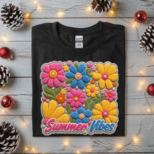 Peut inclure: T-shirt noir orné d'un motif floral coloré avec des fleurs roses, bleues et jaunes, agrémenté de perles et de feuilles vertes. Le texte "Summer Vibes" est brodé. Le t-shirt est présenté sur une surface en bois.