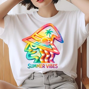 May include: White t-shirt with a vibrant graphic. The design shows a beach scene with a palm tree, sun, and waves, and the words "SUMMER VIBES" in rainbow colours. The shirt is made of a soft material.