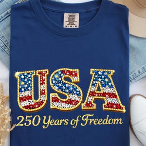 Può includere: T-shirt blu navy con la scritta "USA" in un design scintillante di stelle e strisce rosse, bianche e blu. Il testo "250 Years of Freedom" è sotto la scritta "USA". La maglietta ha uno scollo tondo e l'etichetta del marchio Comfort Colors.