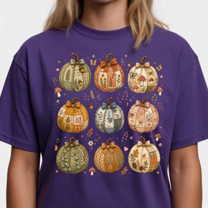 May include: A purple t-shirt featuring a graphic print of nine pumpkins arranged in a grid. Each pumpkin has a unique patchwork pattern with floral, leaf, and mushroom motifs, topped with a small bow and a button at the stem in various colours.