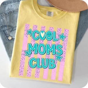 Peut inclure: Un t-shirt jaune avec l'inscription "COOL MOMS CLUB" en turquoise et blanc, orné d'étoiles et d'un cœur. Le motif est placé sur des rayures verticales roses avec un motif moucheté. Le t-shirt est plié sur un jean bleu.