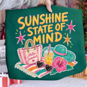 Peut inclure: Un sac fourre-tout vert avec l'inscription "Sunshine State of Mind" en jaune et orange. Le motif comprend un sac de plage, des lunettes de soleil, une boisson, un chapeau et une fleur. Les mots "Hello Summer" sont sur le sac de plage.