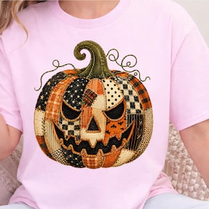 Op de afbeelding: Een lichtroze t-shirt met een grote, centrale afbeelding van een Halloween-pompoen. De pompoen is ontworpen met een patchworkpatroon van verschillende stoffen in oranje, zwart en wit, waaronder patronen met sterren, stippen en ruiten.