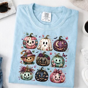May include: A light blue crew neck t-shirt featuring a graphic print of nine stylised pumpkins and a ghost. The pumpkins are decorated with various patterns, flowers, and colours including pink, purple, black, and teal. The tag reads Comfort Colors.