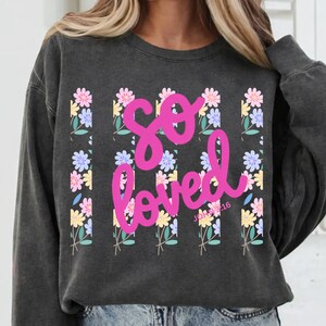 Peut inclure: Sweat-shirt gris foncé avec l'inscription "so loved" en rose vif. Le motif comprend des rangées verticales de fleurs colorées roses, bleues et jaunes. Le texte "John 3:16" est en dessous du mot "loved".