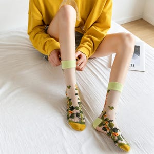 May include: Sheer socks with a floral pattern. The socks have a yellow toe and heel, a light green cuff, and a design of green leaves and yellow flowers. The socks are worn by a person sitting on a white bed.